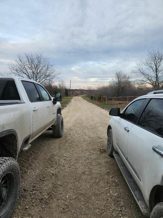 Two white vehicles parked on a gravel road with trees on both sides under a cloudy sky.