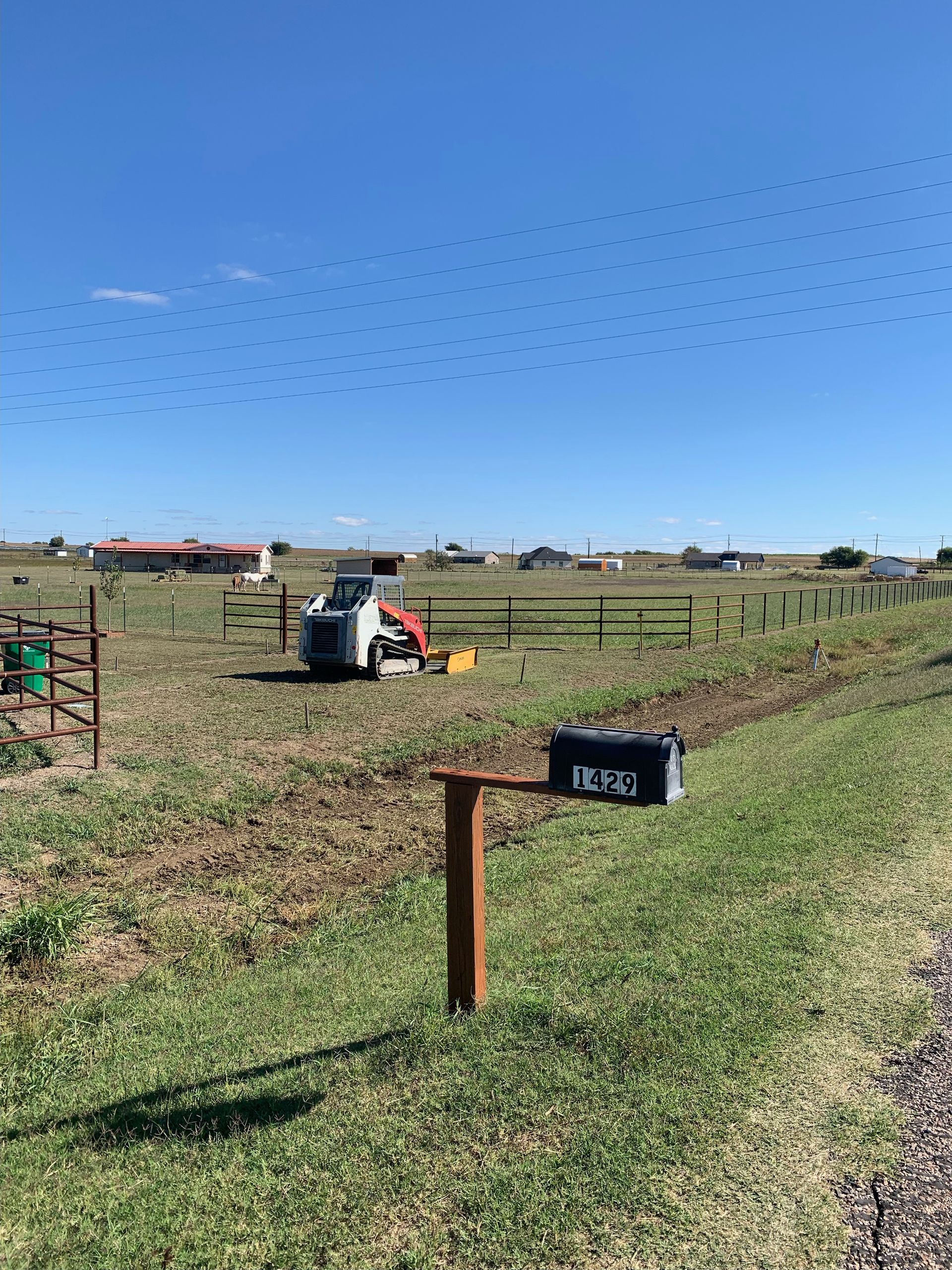 Field with a small tractor, mailbox, and buildings under a blue sky, with small, dark objects seemingly in the air.