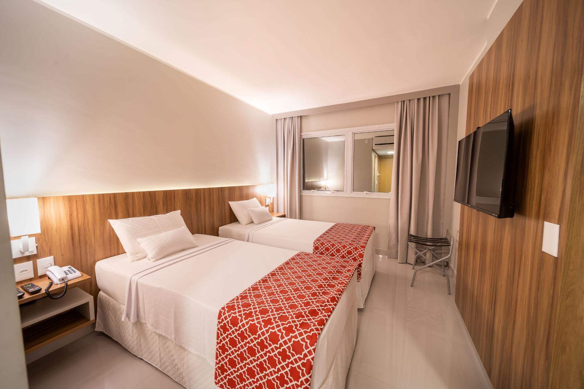 Duas camas de hotel com passadeiras estampadas em vermelho e branco, TV na parede com painéis de madeira.