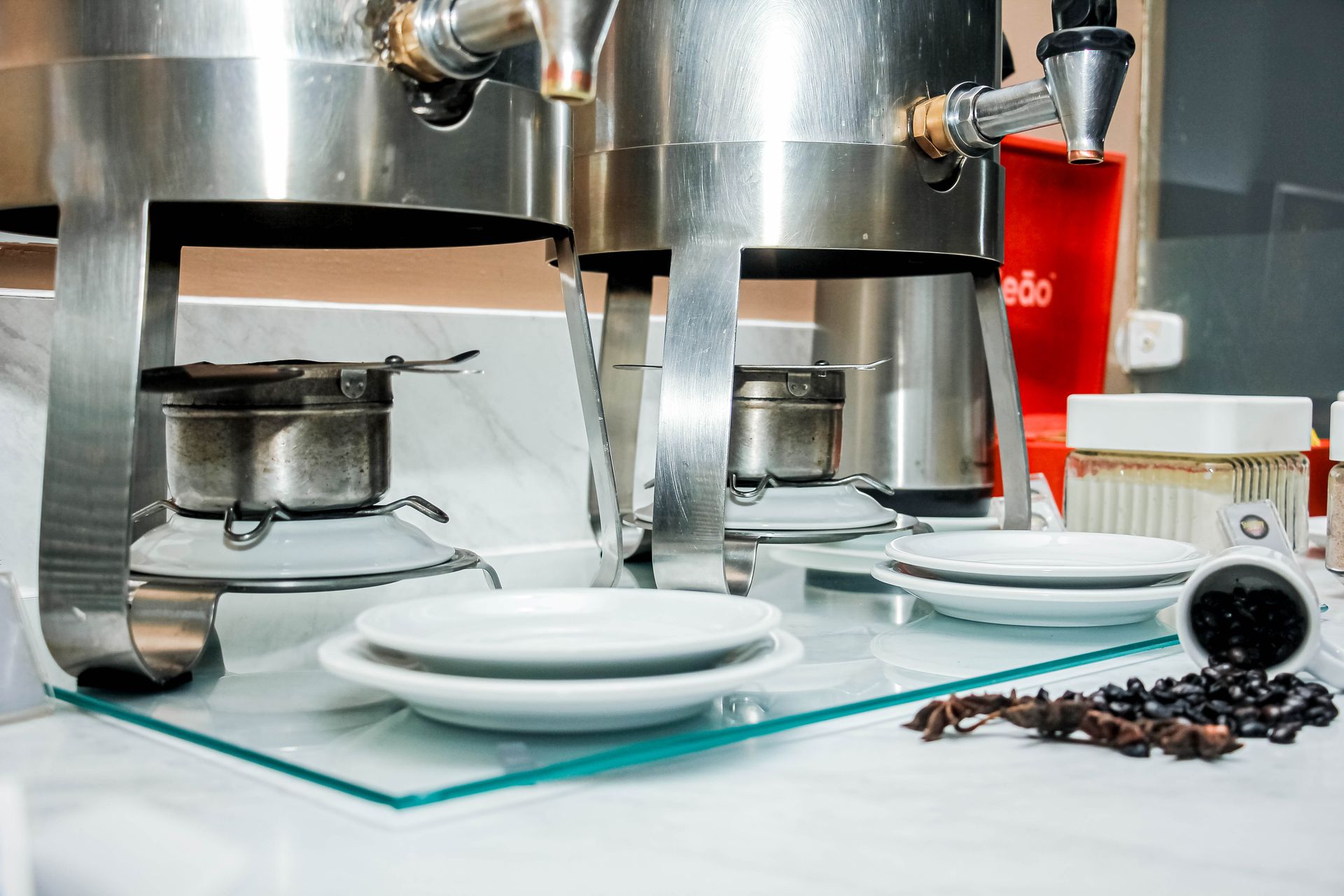 Cafeteiras com queimadores de combustível, pratos empilhados, grãos de café e xícaras sobre uma superfície de vidro.