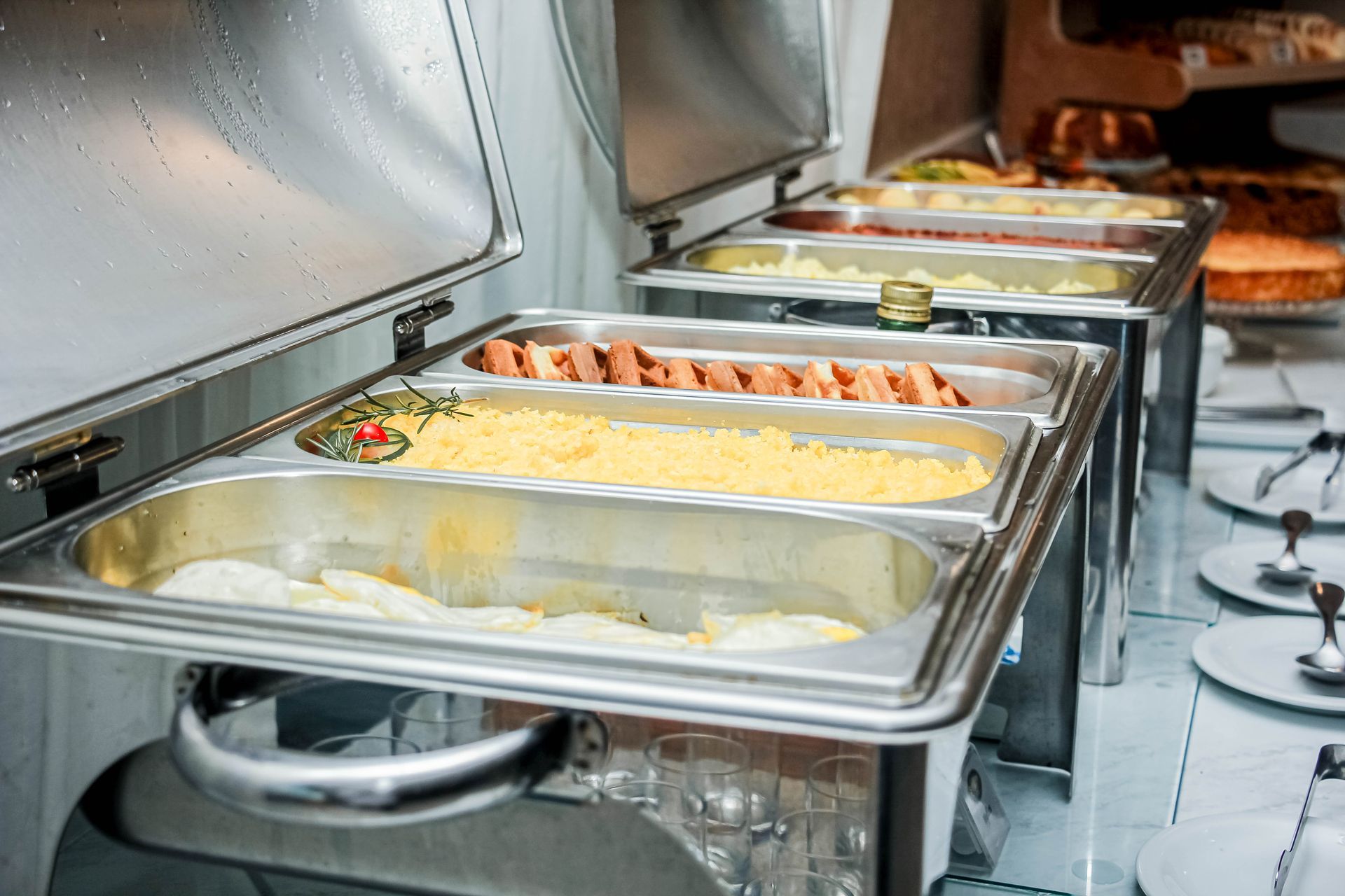 Estação de comida tipo bufê com bandejas aquecidas contendo vários itens de comida cozida, com pratos brancos ao lado.