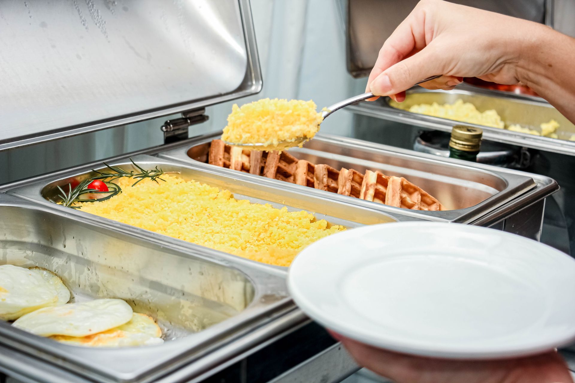 Pessoa servindo comida de um bufê com aquecedores de comida de metal; segurando um prato e uma colher de servir.
