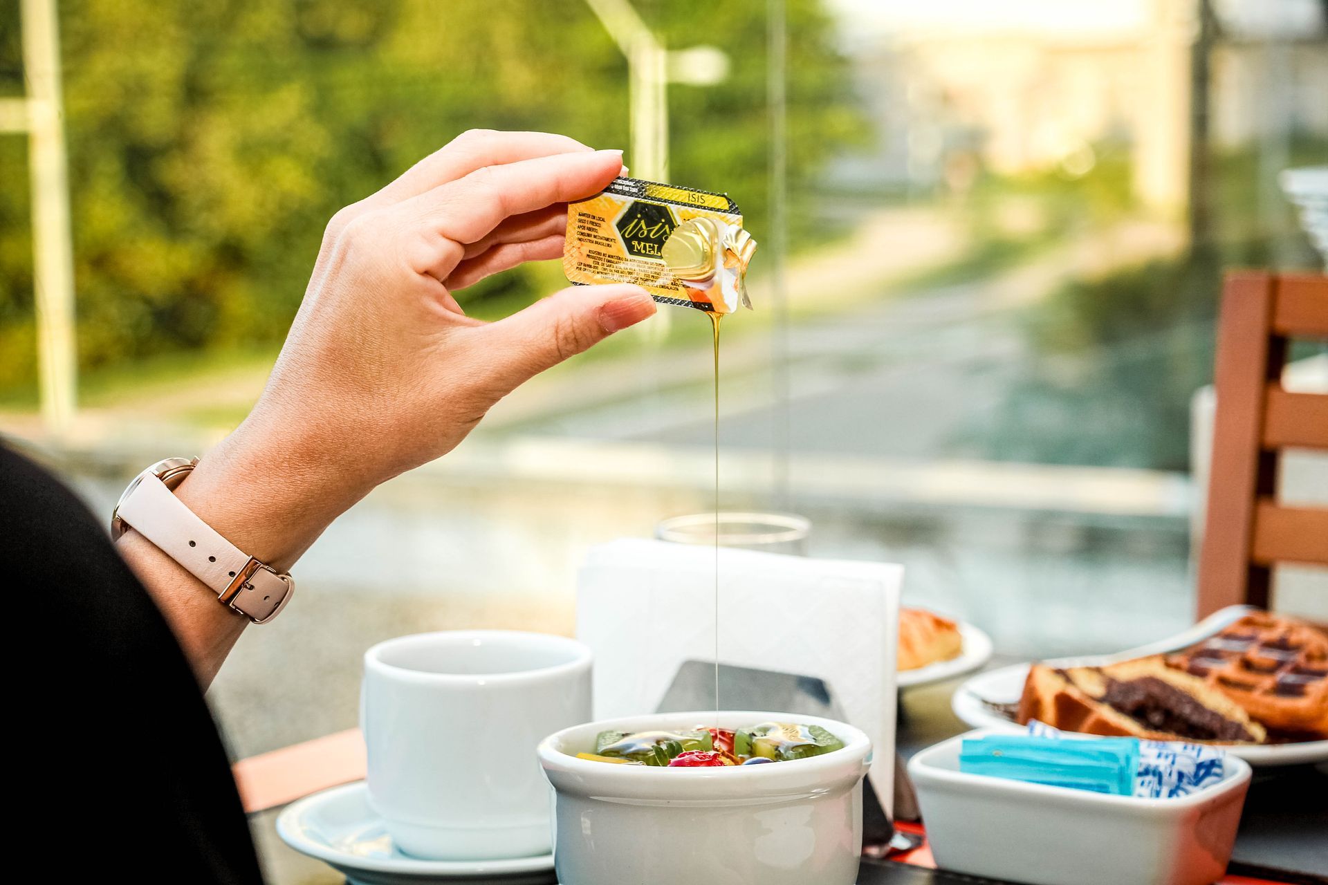 Mão segurando um pastel quadrado com calda pingando sobre uma mesa posta com itens de café da manhã.