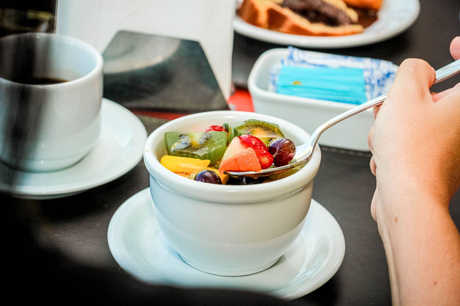 Pessoa comendo salada de frutas em uma tigela branca com café e um doce ao fundo.
