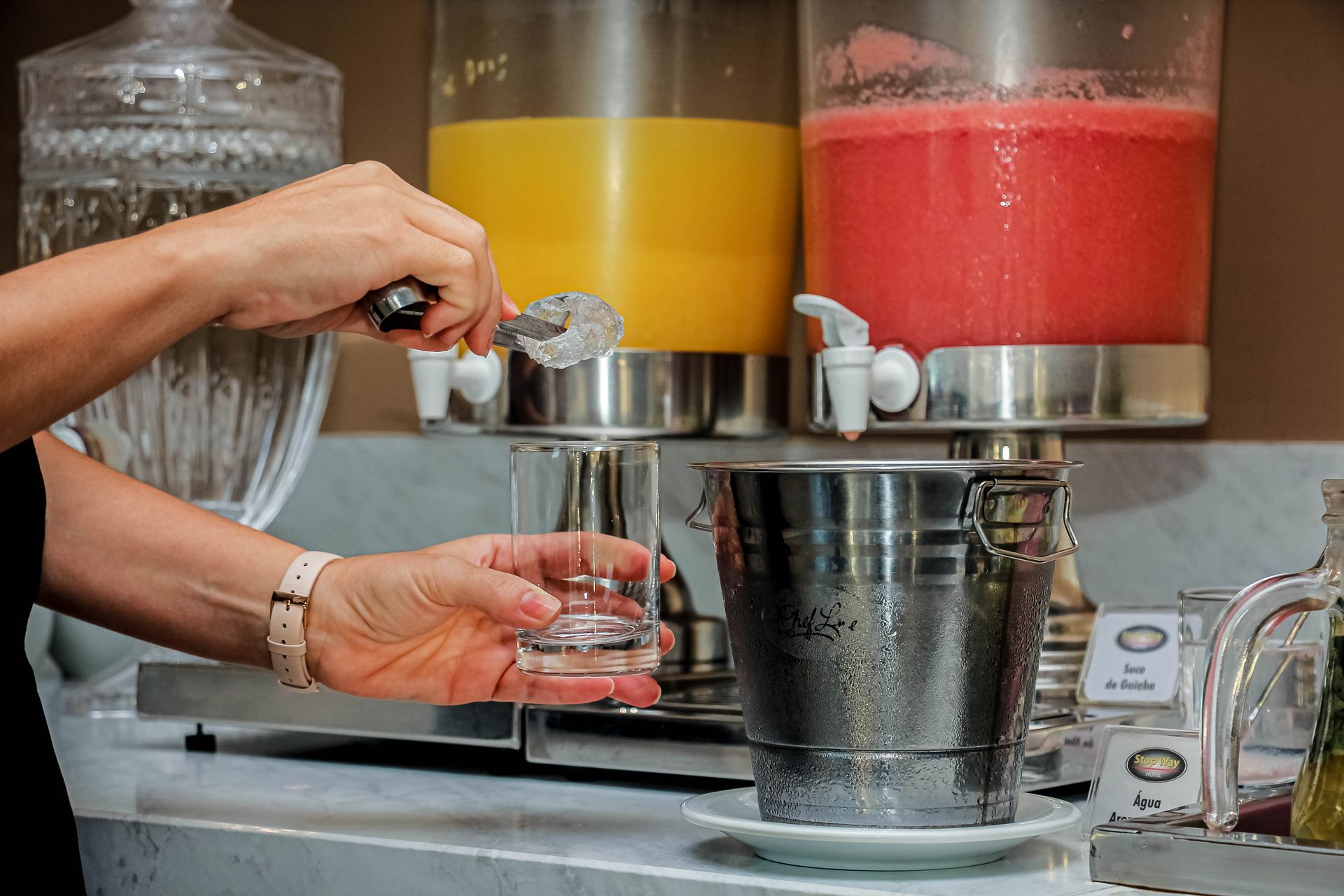 Pessoa colocando gelo em um copo, com dispensadores de suco de laranja e vermelho ao fundo.