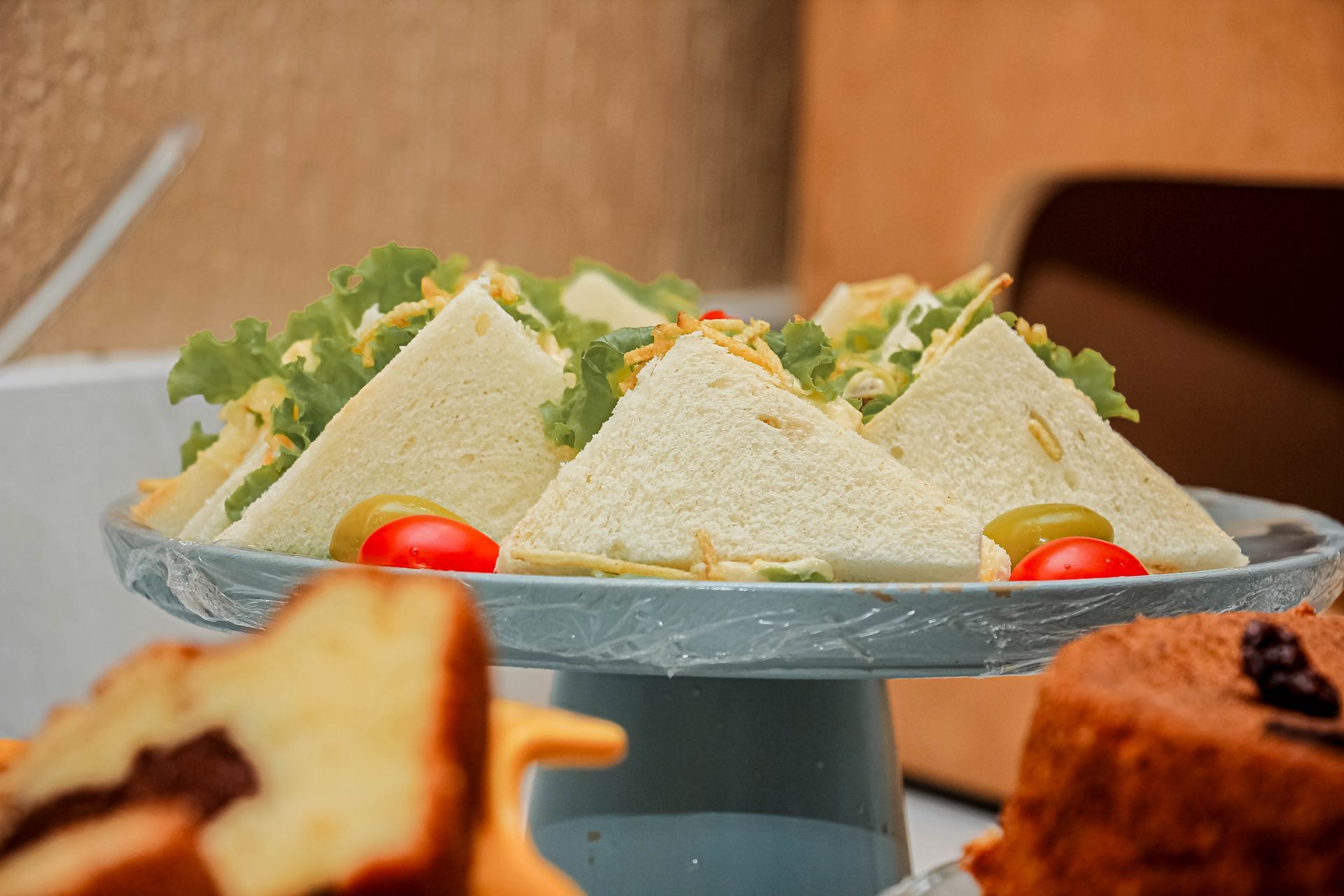 Sanduíches triangulares em um suporte de bolo azul com tomates, azeitonas e uma fatia de bolo em primeiro plano.
