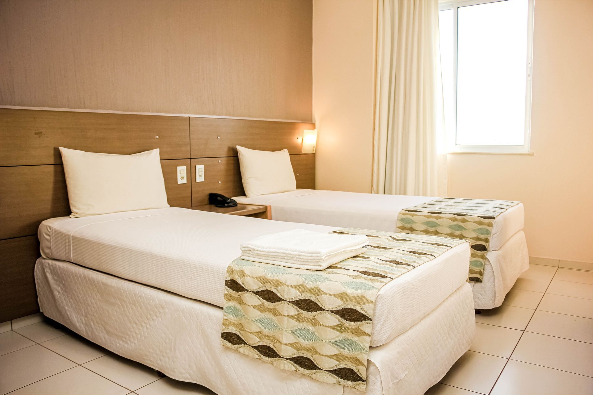 Duas camas de solteiro em um quarto de hotel bege, com cortinas estampadas e uma janela com cortinas brancas.