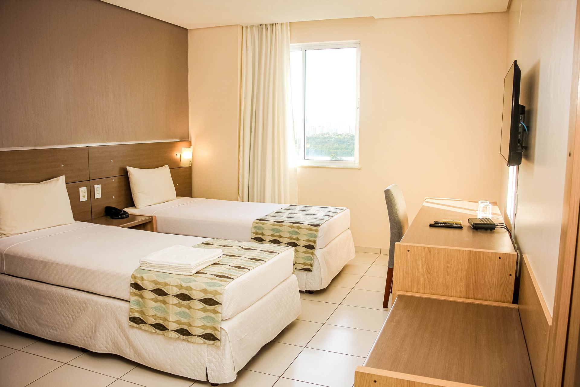 Quarto de hotel com duas camas de solteiro, escrivaninha, janela e TV na parede. Paredes bege, decoração em tons neutros.