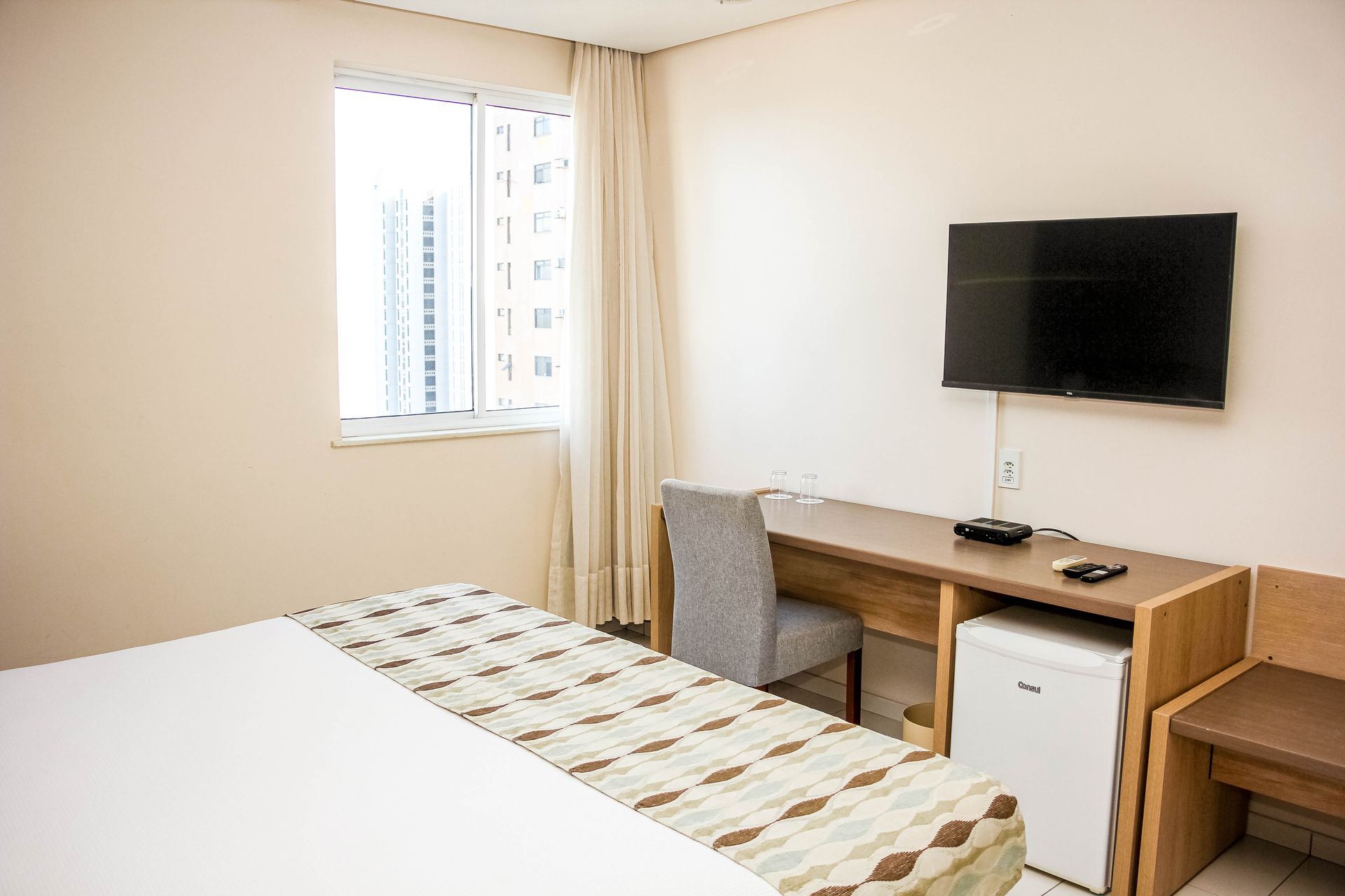 Quarto de hotel: cama, escrivaninha com cadeira, TV, janela e frigobar. Paredes e cortinas bege.