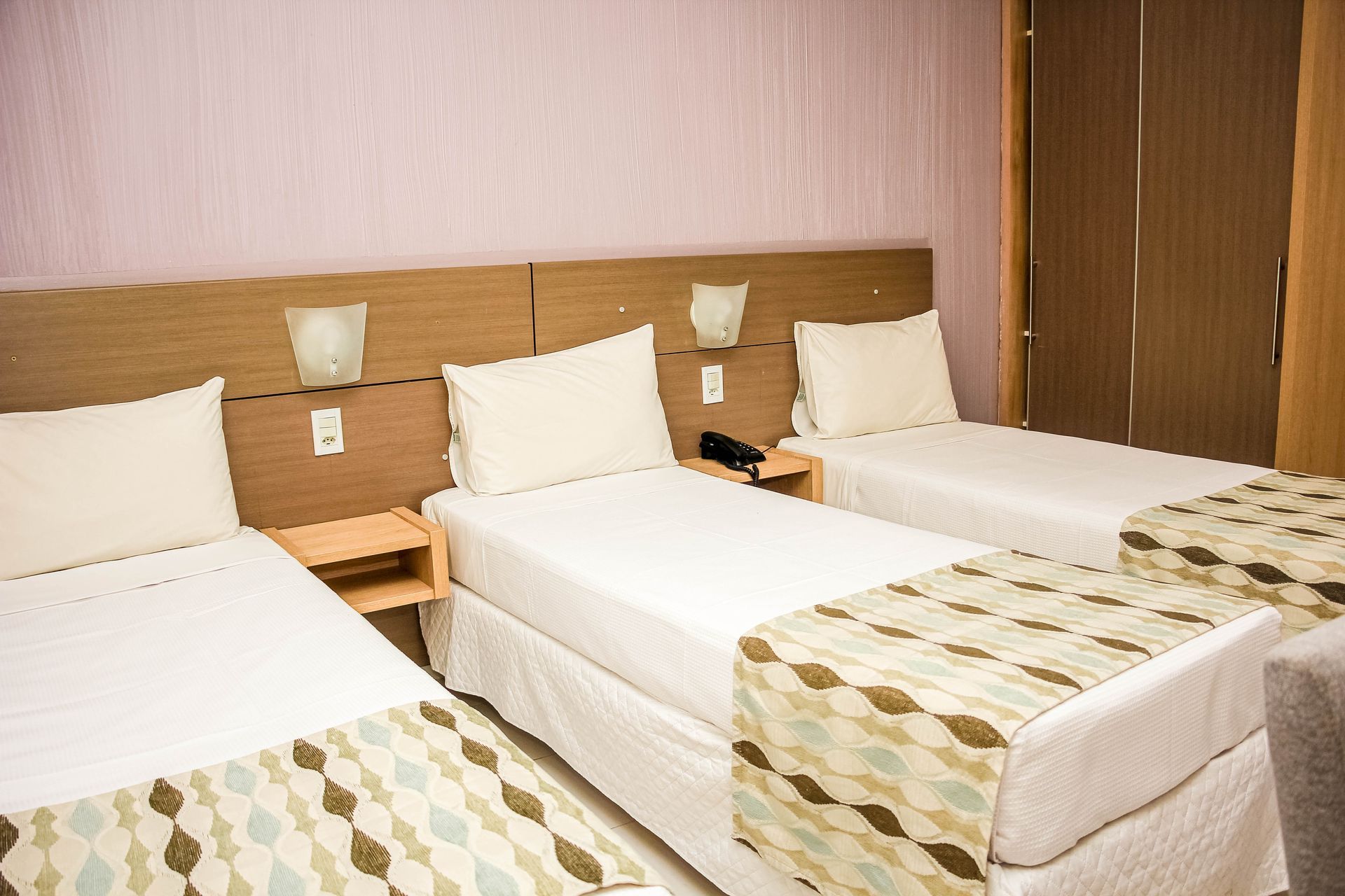 Três camas em um quarto de hotel com lençóis brancos, cabeceiras de madeira e passadeiras estampadas.