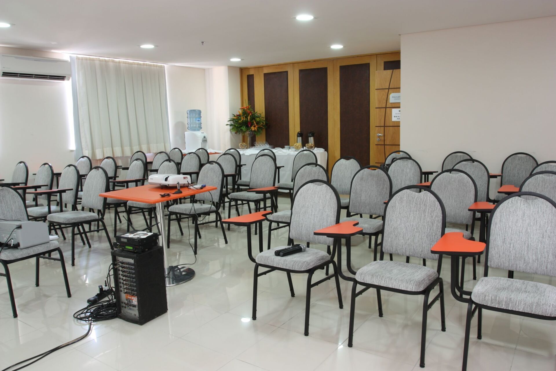 Sala de aula vazia com fileiras de cadeiras cinzas, projetor sobre a mesa e um sistema de alto-falantes.