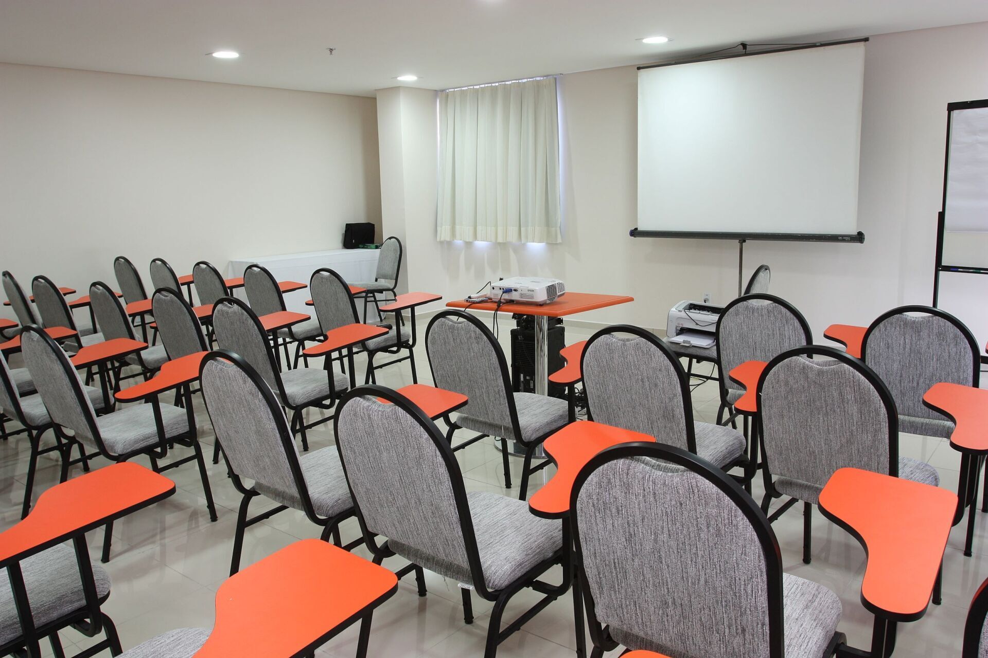 Sala de aula com fileiras de cadeiras com braços de tablet laranja, tela de projeção e projetor.