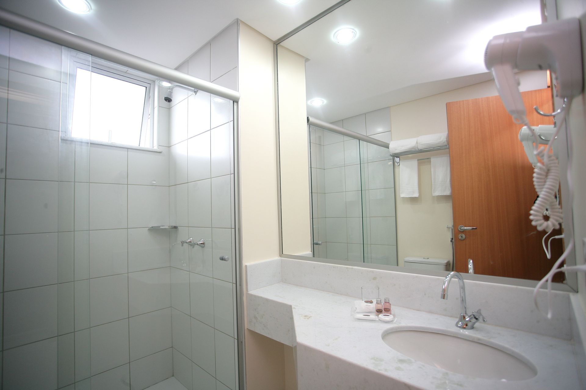 Banheiro com chuveiro de azulejos brancos, bancada de mármore e um espelho grande.