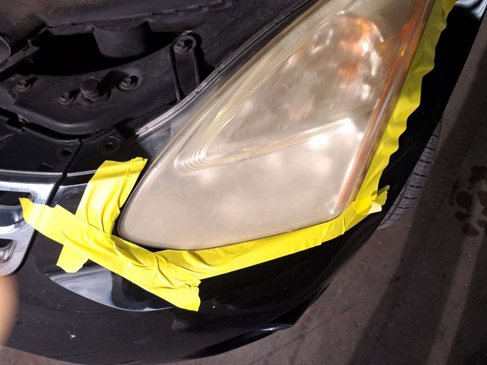 A car headlight prepped for restoration with yellow masking tape applied to the surrounding bumper and hood edges.