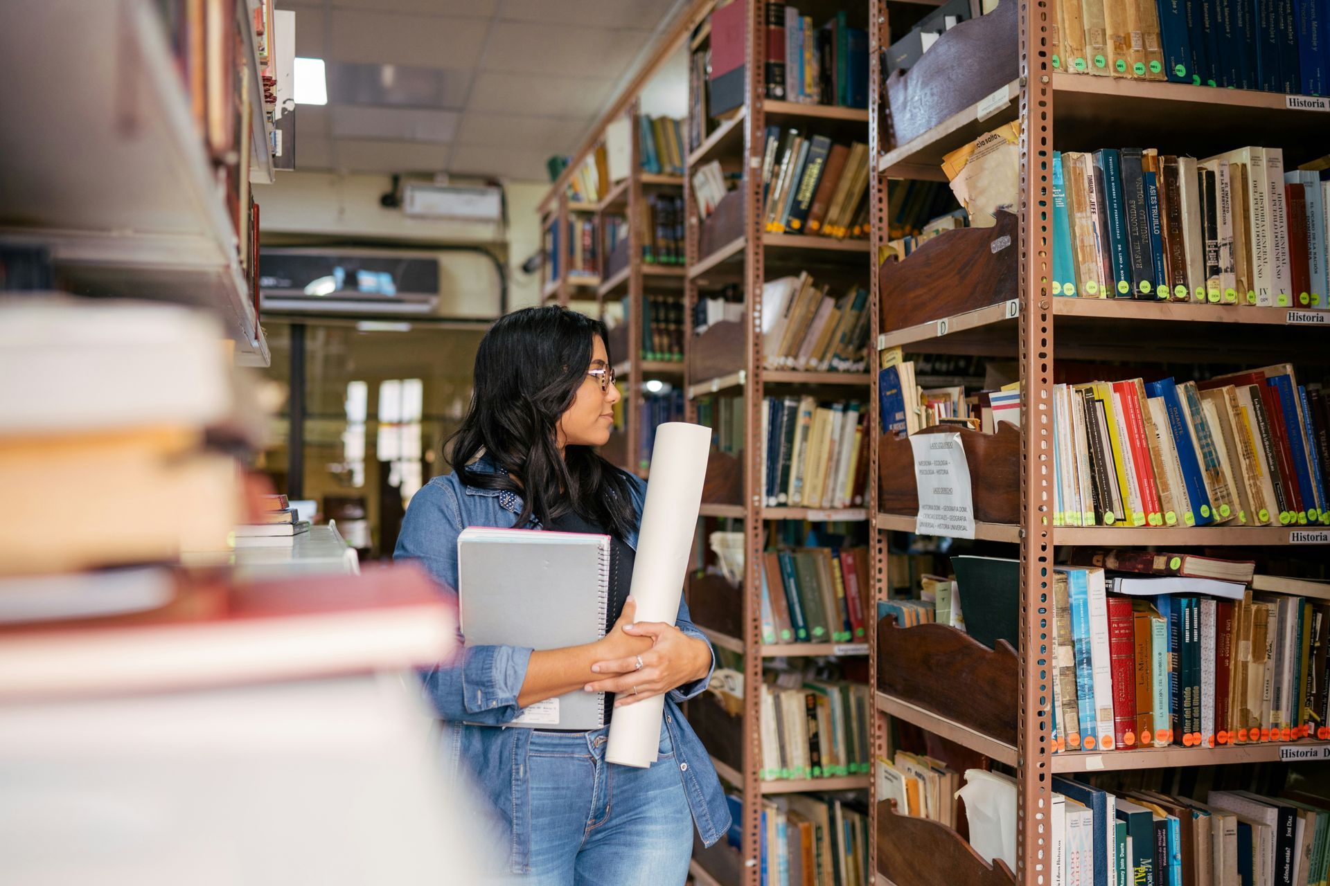 A woman is standing in a library holding a laptop and a piece of paper.