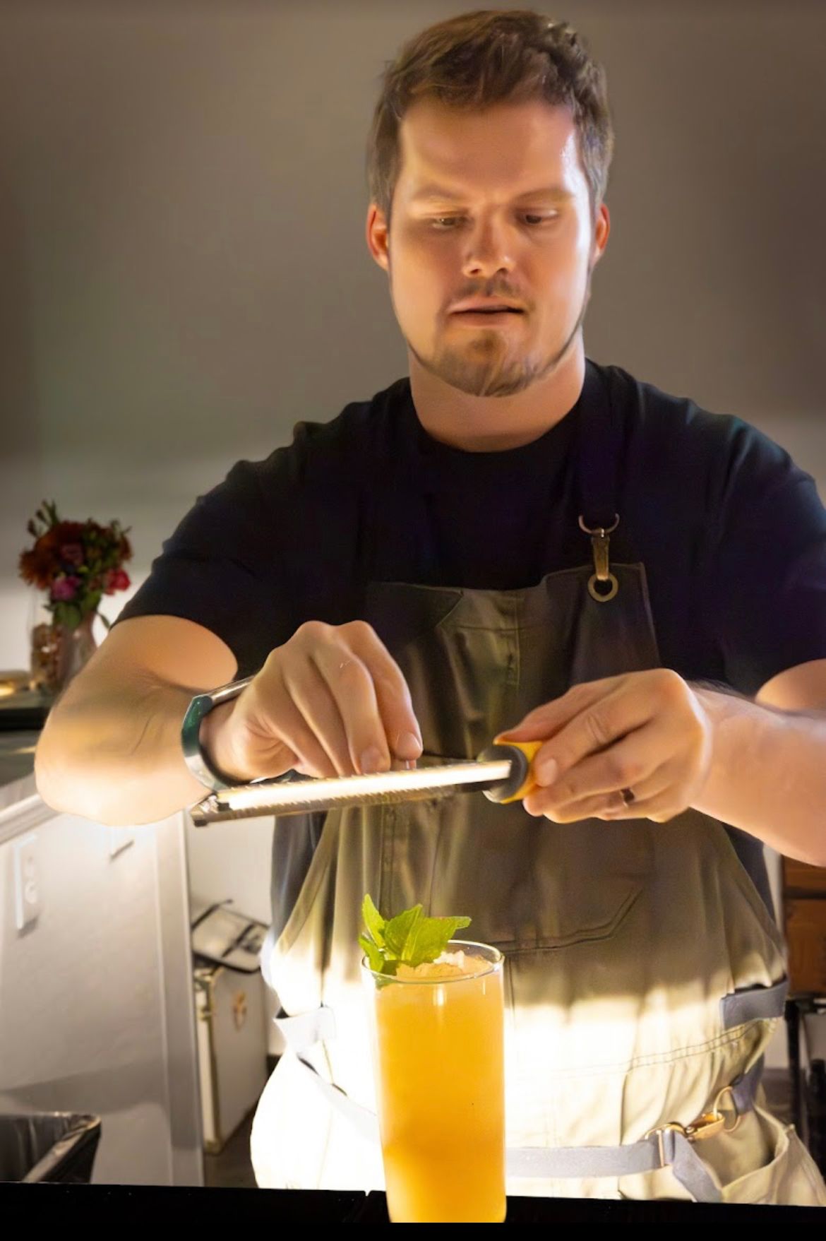 Bartender zesting orange peel over a tall cocktail. He wears a black shirt and apron. Indoor setting.