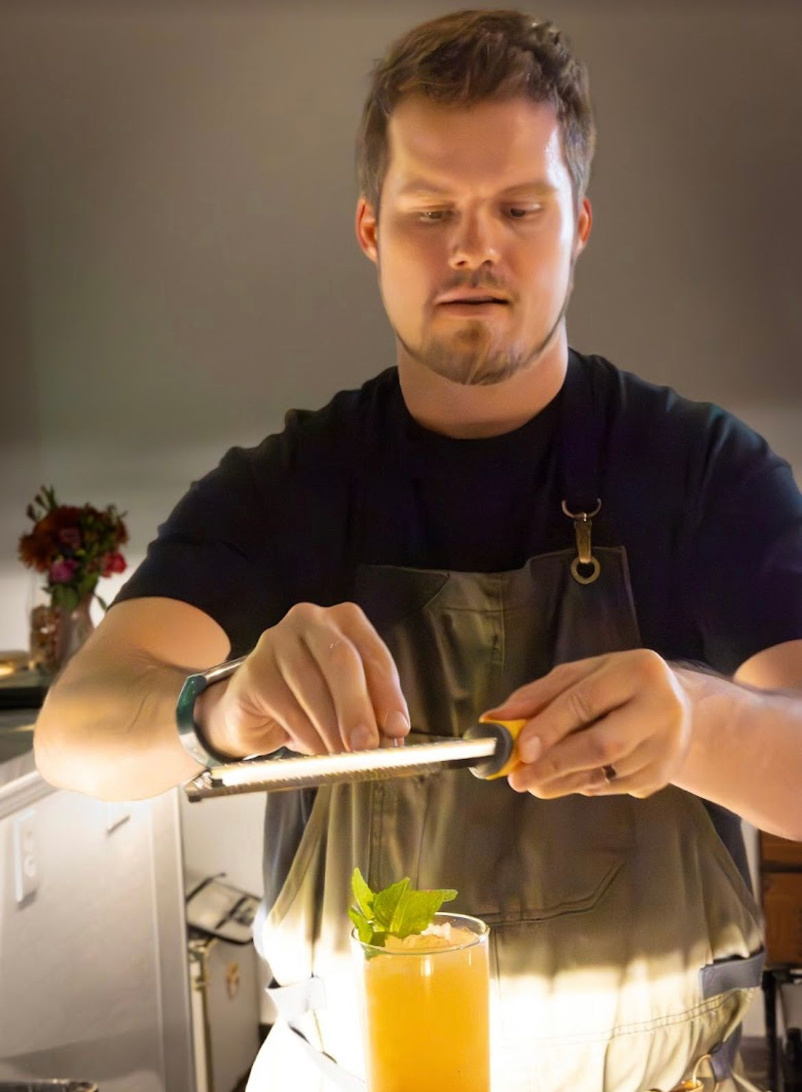 Bartender zesting orange peel over a cocktail. He's wearing a black shirt and apron. Indoor setting.