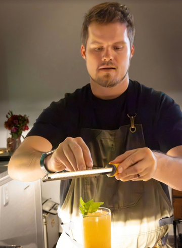 Bartender zesting orange peel over a cocktail. He's wearing a black shirt and apron. Indoor setting.