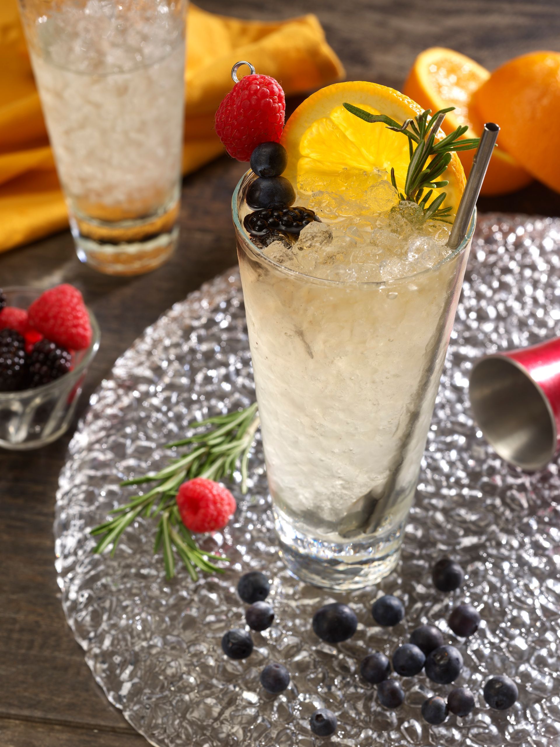 Cocktail with berries, orange slice, rosemary, crushed ice, and a stainless steel straw on a glass platter.