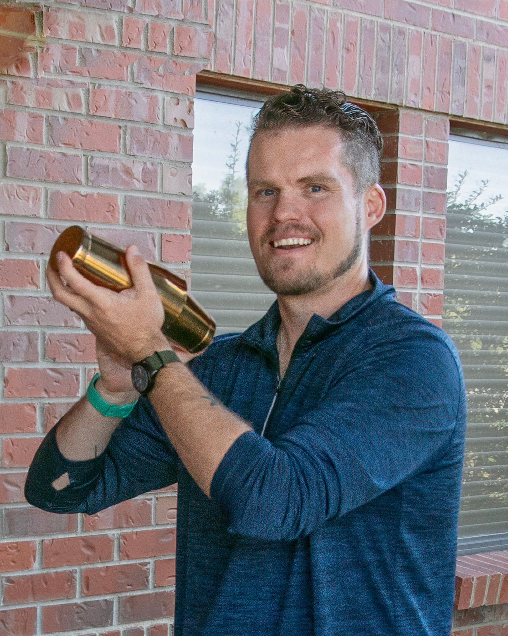 Make It a Double shaking a cocktail shaker, smiling, wearing a blue sweater, standing against a brick wall.