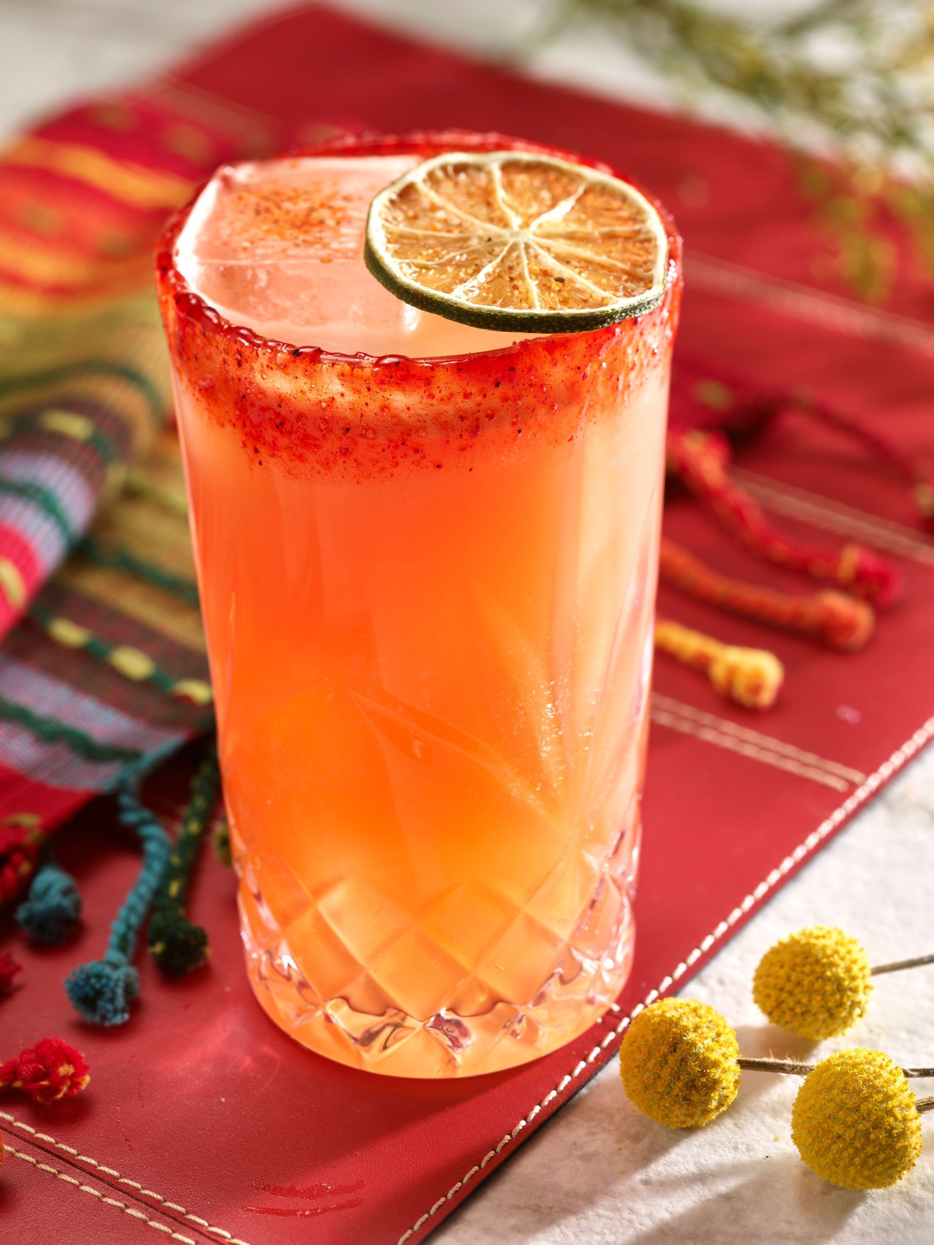 Tall cocktail with red chili rim and lime garnish, on a red cloth.