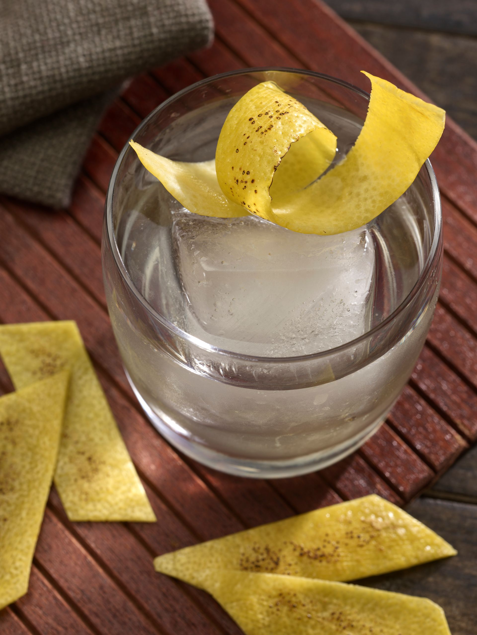 Cocktail in a clear glass with a large ice cube, garnished with lemon peel on a wooden surface.