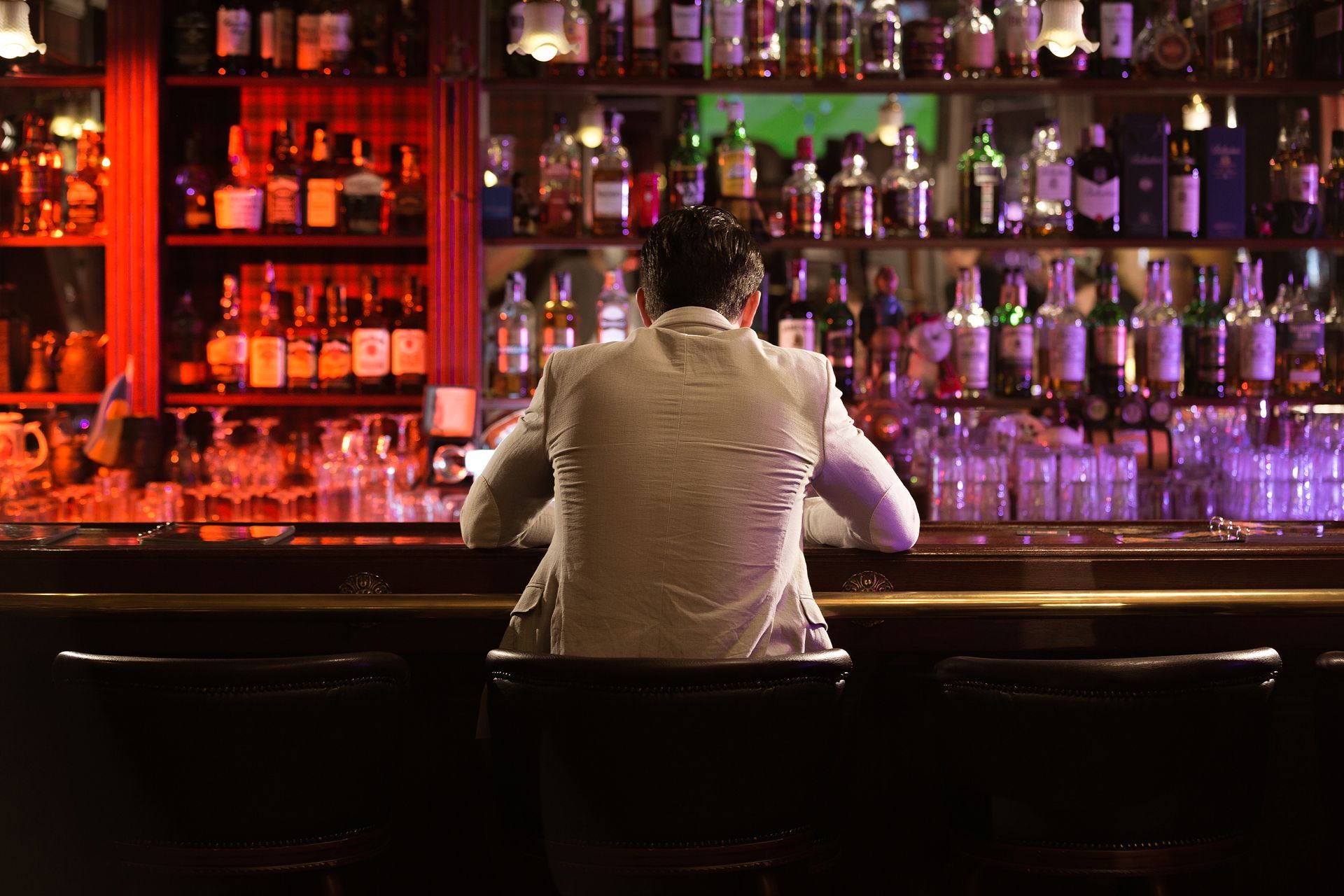 Man in a white suit sits alone at a bar, looking at bottles behind the counter.