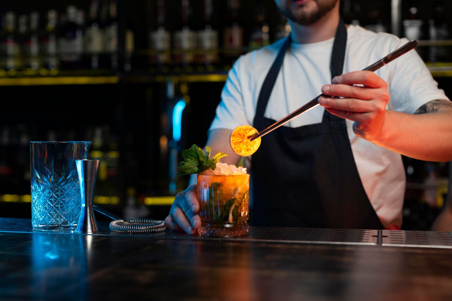 Bartender garnishes cocktail with orange slice using a long stick; bar setting.