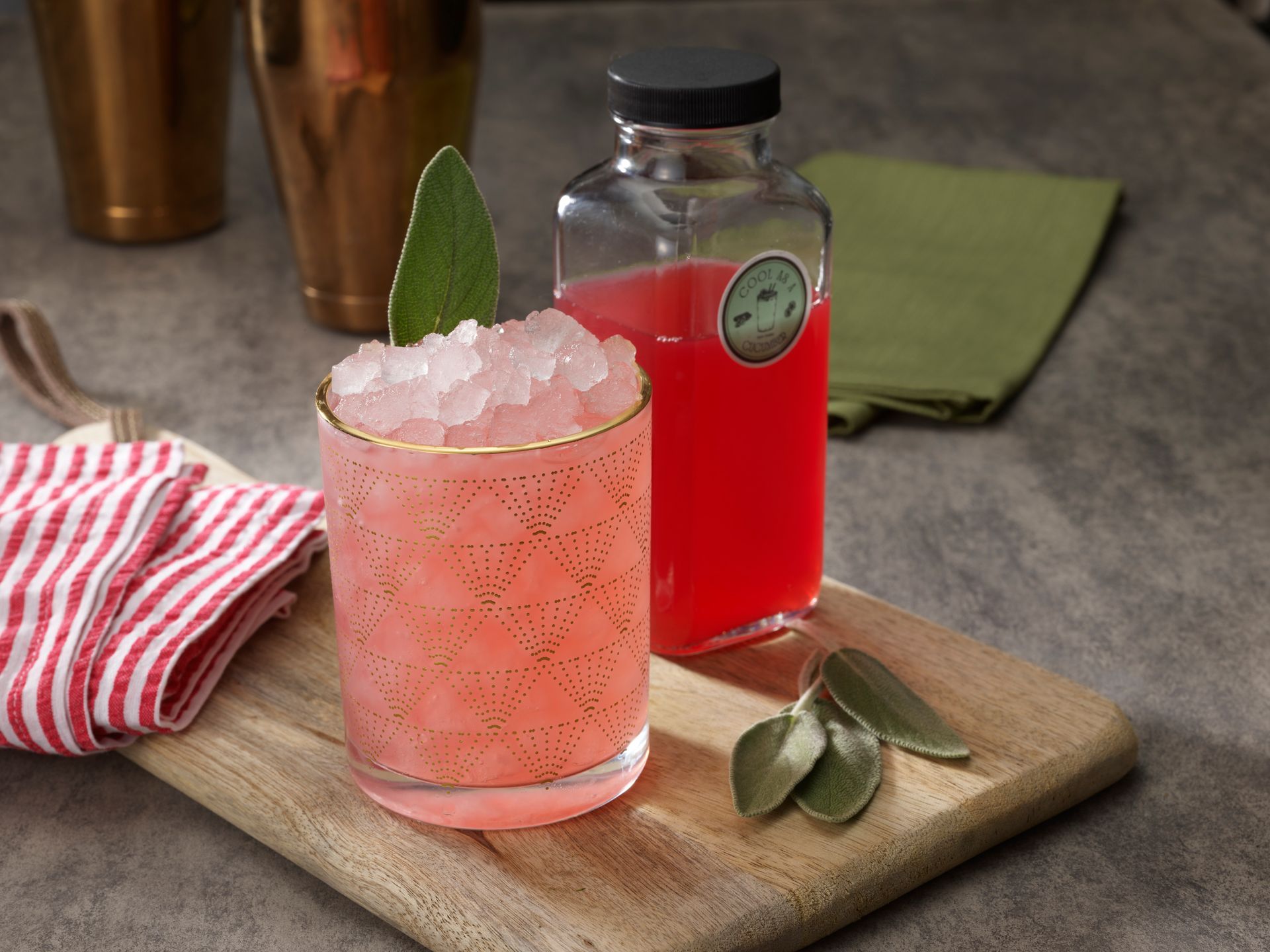 Cocktail with pink drink and crushed ice, sage garnish, bottle of red liquid, wooden board, and gold bar tools.