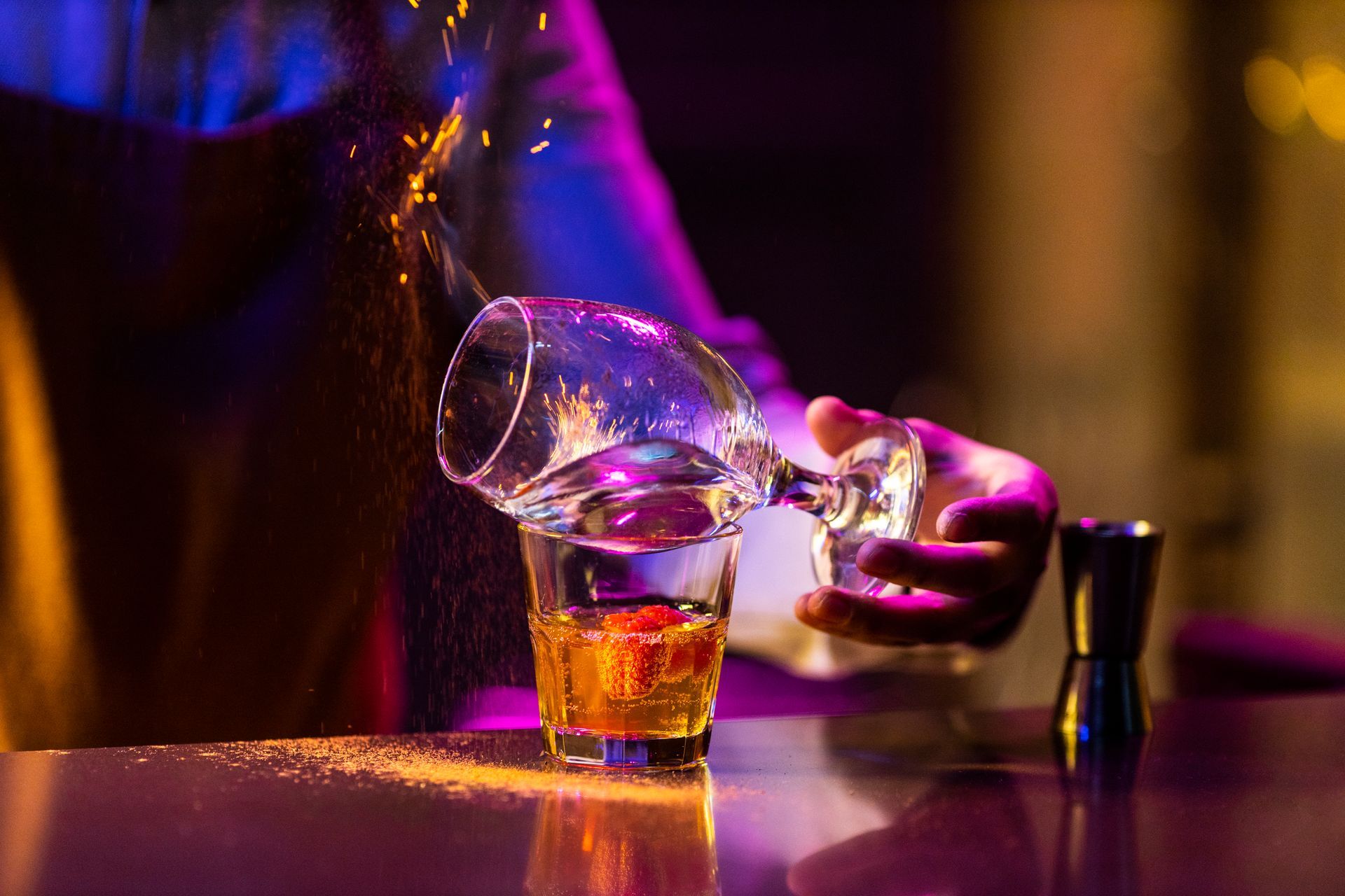 Bartender pouring drink, shot glass with liquid, spilled sugar, bar setting with colored lights.
