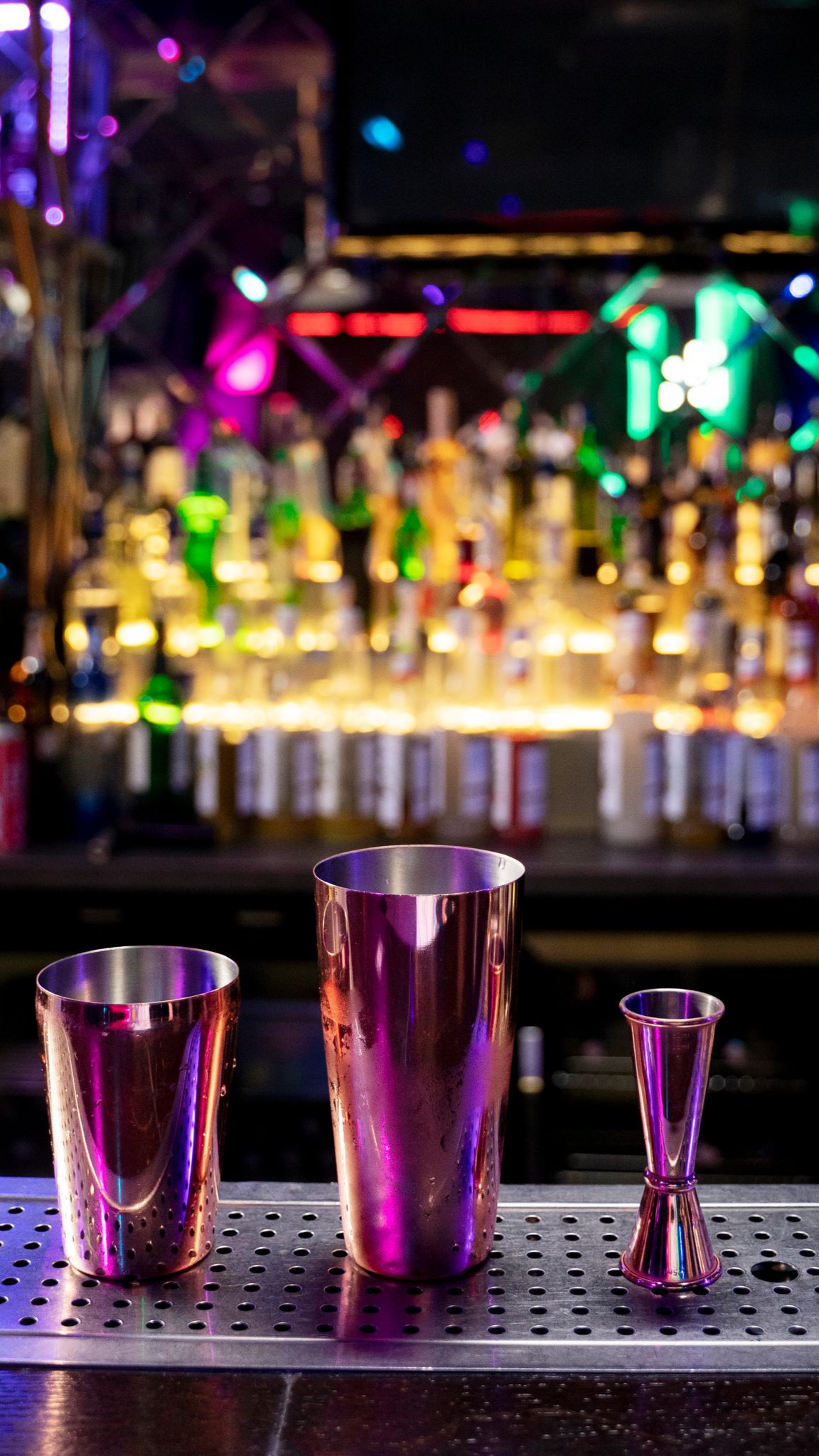 Copper cocktail shaker, mixing glass, and jigger on bar in a brightly lit bar.