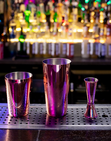 Three copper bar tools on a bar: a cocktail shaker, a mixing glass, and a jigger. Background of colorful bottles.
