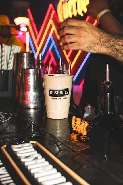 Bartender zesting orange peel over a cocktail. He's wearing a black shirt and apron. Indoor setting.