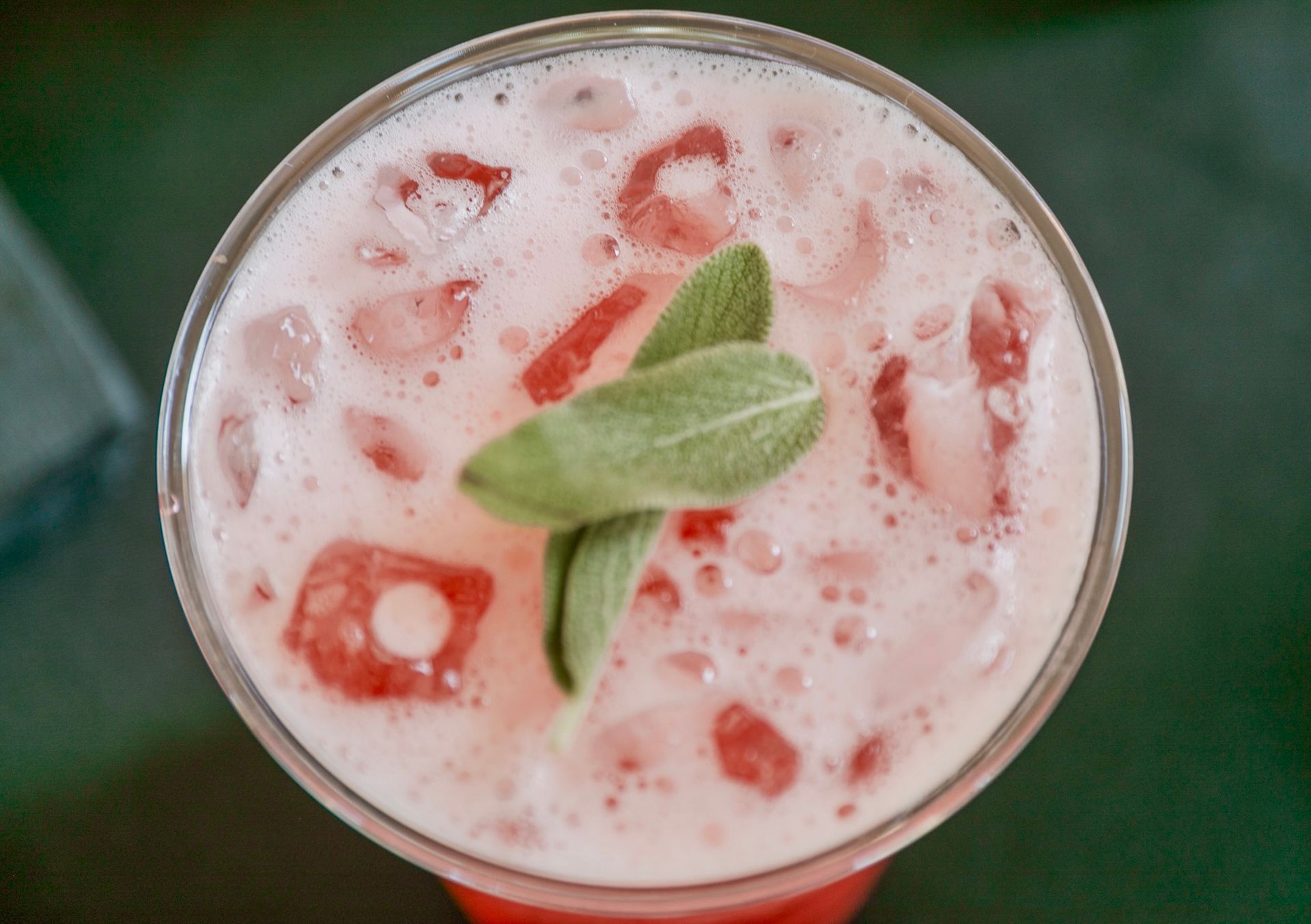 Overhead view of a pink cocktail with ice, garnished with green sage leaves, in a clear glass.