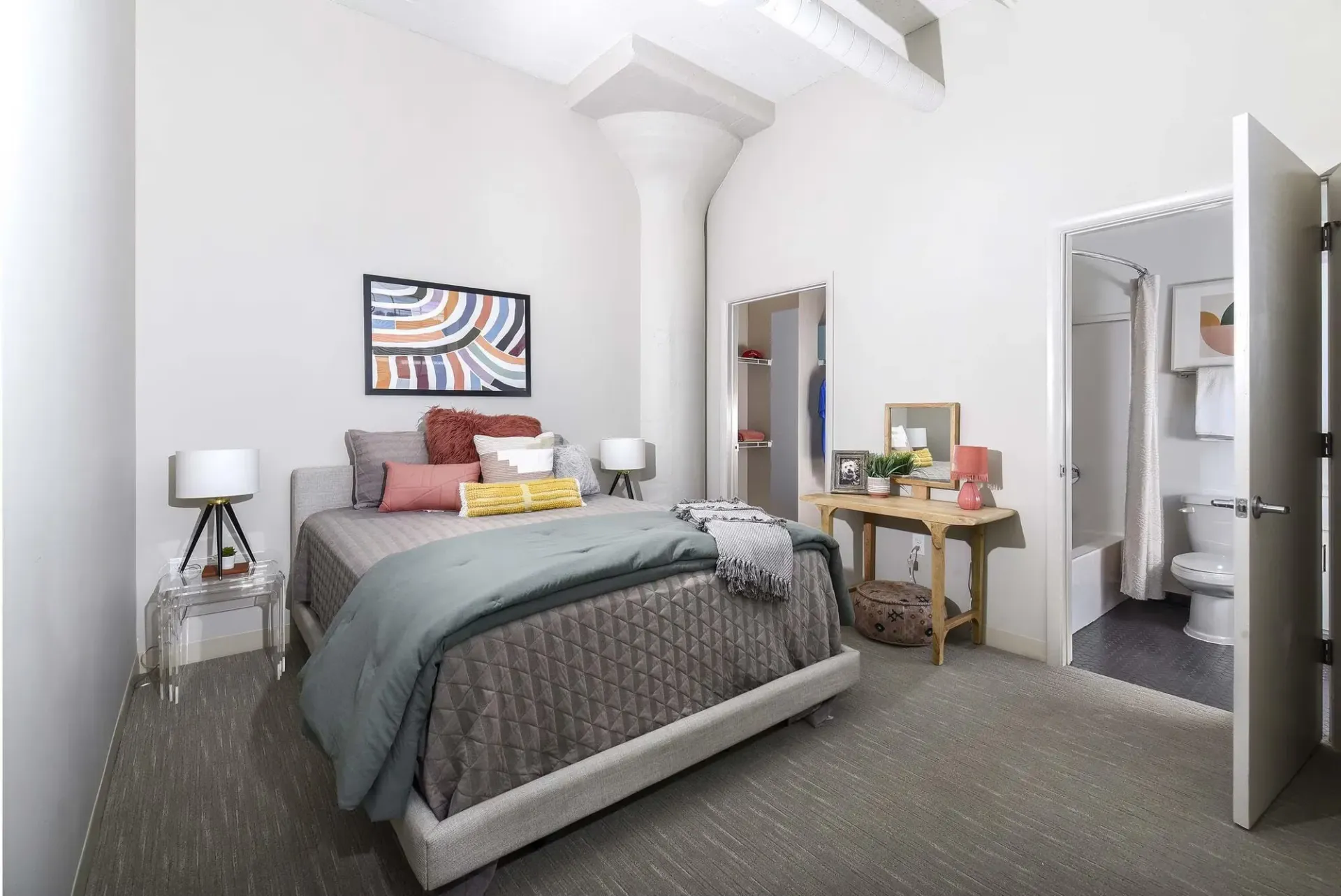 Furnished bedroom with a gray upholstered bed, two nightstands, open closet, and adjoining bathroom at Terminal 21, a premier apartment community in Downtown Pittsburgh, Pennsylvania.