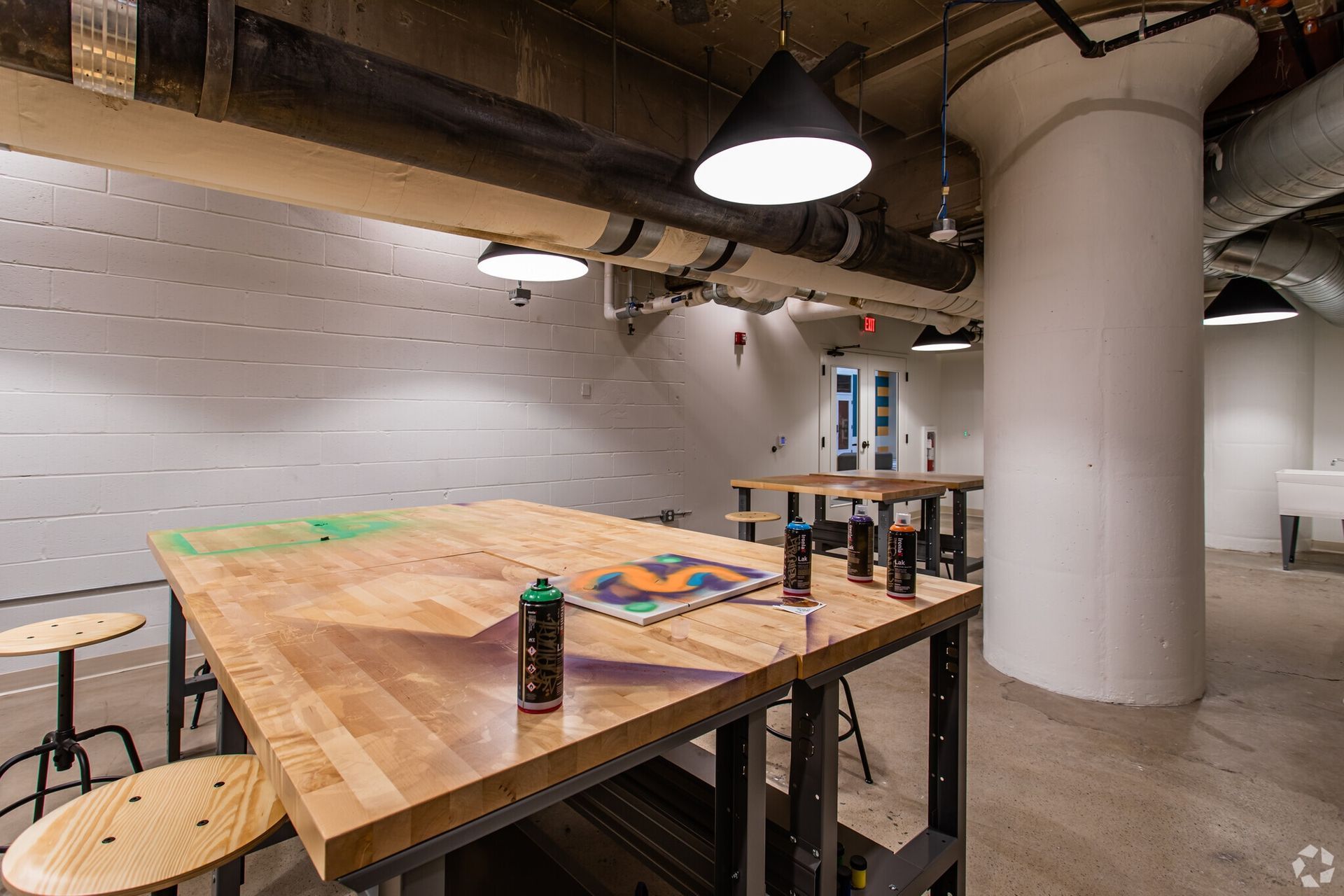 Industrial workshop area with large wooden tables, stools, and exposed pipes at Terminal 21, a premier apartment community in Downtown Pittsburgh, Pennsylvania.