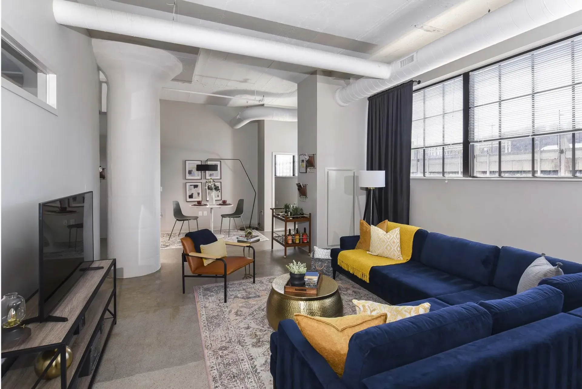 Industrial-style living room with a navy blue sectional, yellow pillows, and exposed ducts at Terminal 21, a premier apartment community in Downtown Pittsburgh, Pennsylvania.
