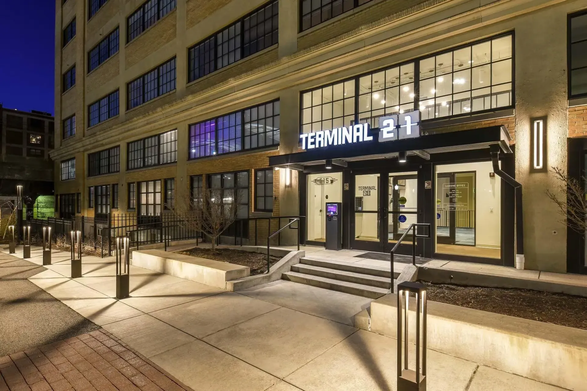 Night exterior view of a modern apartment building entrance with Terminal 21 sign and lit pathway at Terminal 21, a premier apartment community in Downtown Pittsburgh, Pennsylvania.
