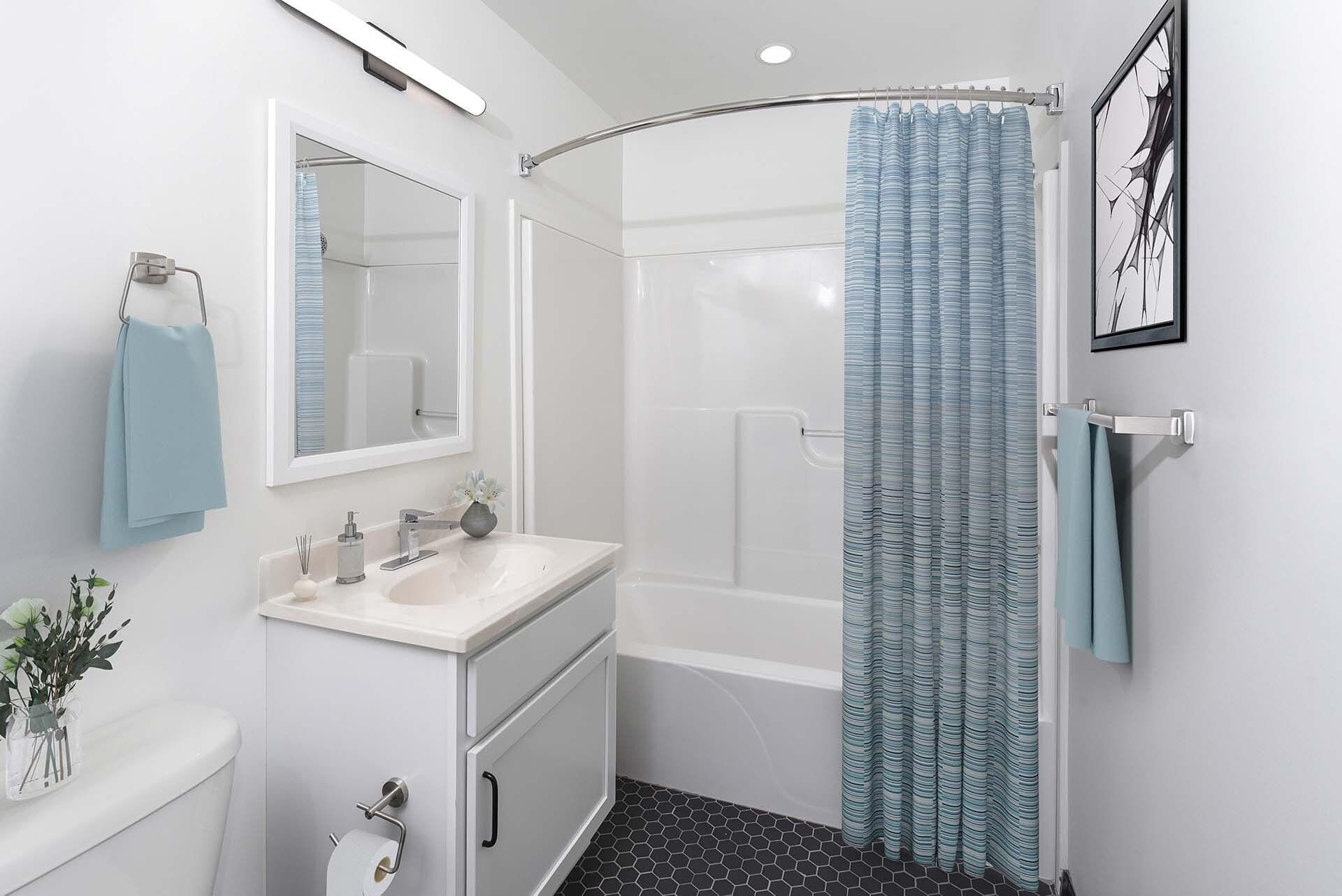 Modern apartment bathroom with vanity, mirror, toilet, and tub/shower with curved rod and blue curtain at Terminal 21, a premier apartment community in Downtown Pittsburgh, Pennsylvania.