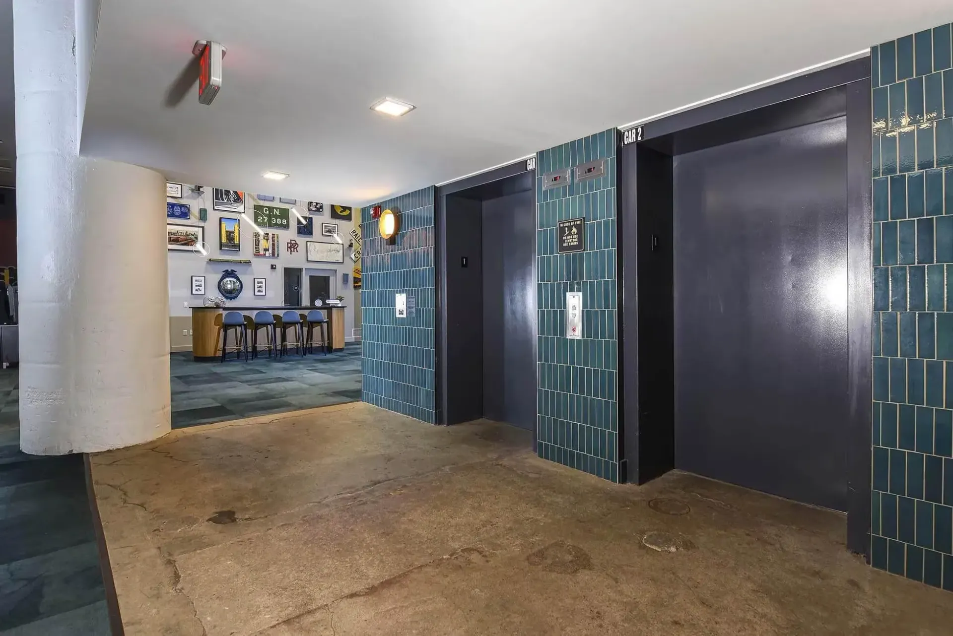 Elevator lobby with teal tile walls, two elevator doors, a concrete column, and a bar area in the background at Terminal 21, a premier apartment community in Downtown Pittsburgh, Pennsylvania.