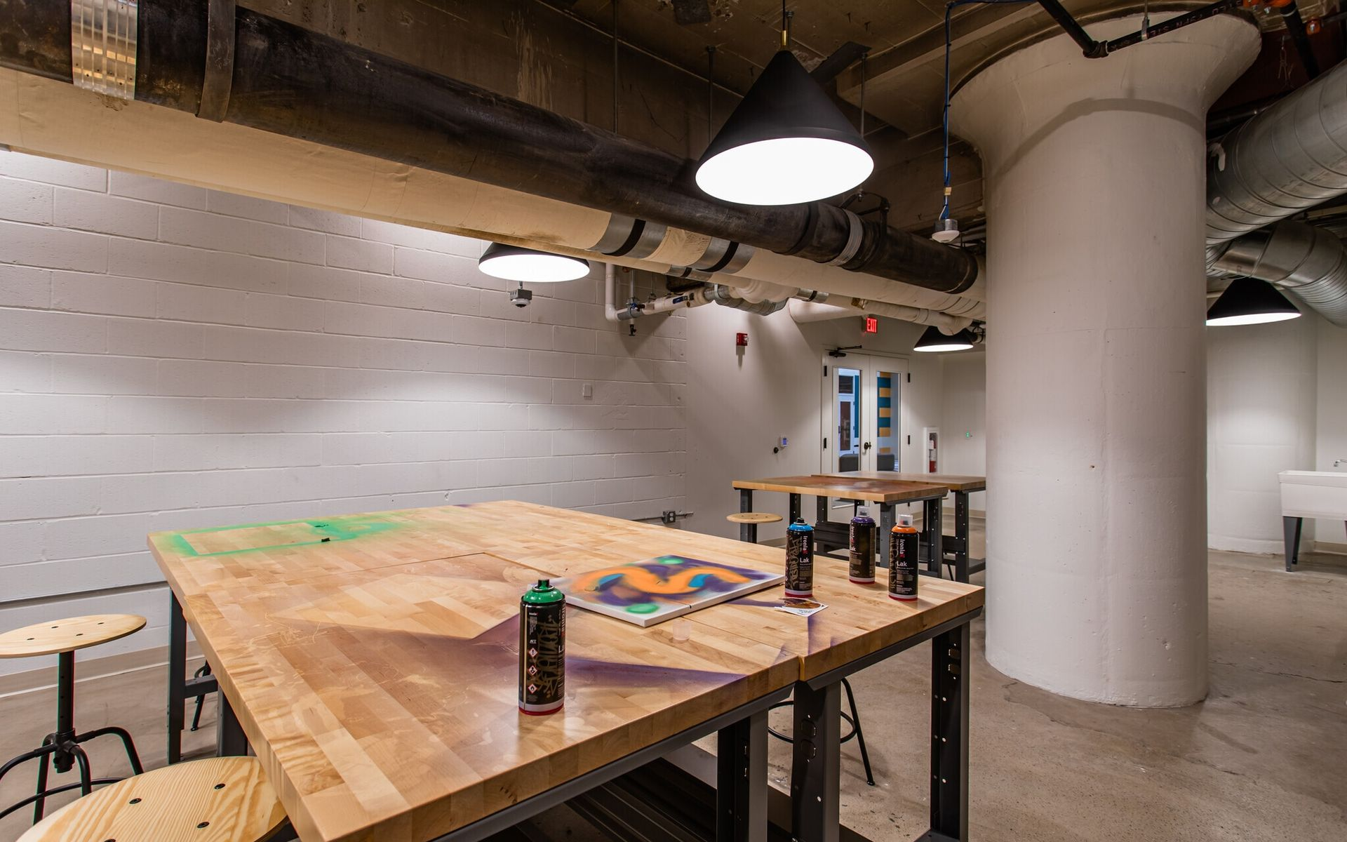 Industrial workshop area with large wooden tables, stools, and exposed pipes at Terminal 21, a premier apartment community in Downtown Pittsburgh, Pennsylvania.