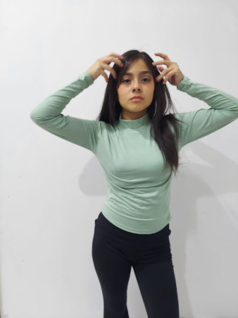 Mujer de cabello oscuro con una camisa verde menta y pantalones negros, con las manos levantadas cerca de la cabeza, contra una pared blanca.