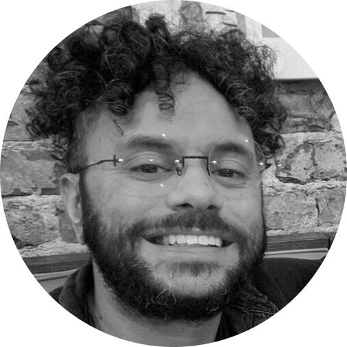 Man with curly hair, glasses, and beard smiles in front of a brick wall.
