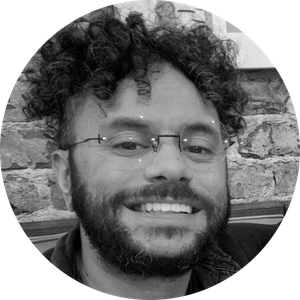 Smiling man with glasses and curly hair, in front of a brick wall. He has a beard and appears to be indoors.