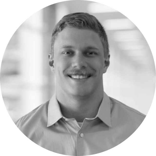 Man with mustache smiles, wearing a light-colored button-down shirt in a blurred office setting.