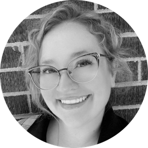 Smiling woman with curly blonde hair and glasses in front of a brick wall.