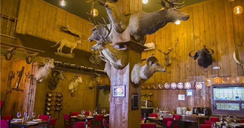 Interior del restaurante con cabezas de animales disecadas en las paredes de madera. Mesas de comedor con sillas rojas.