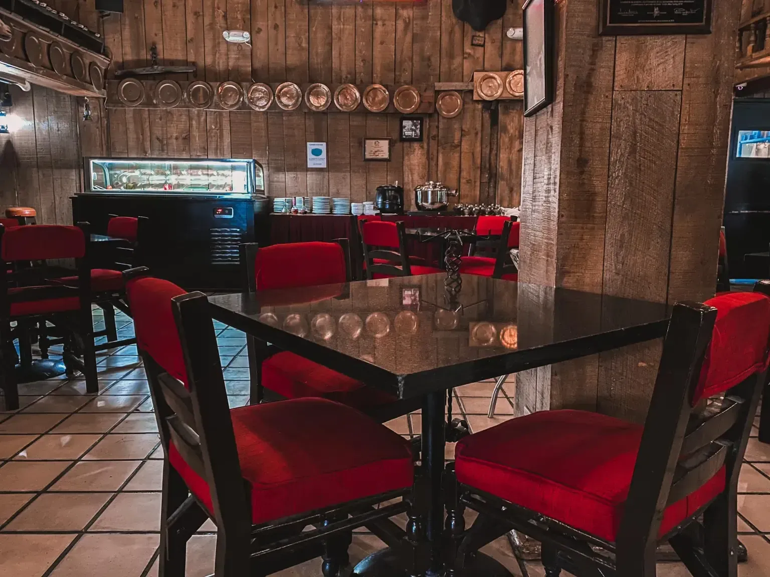 Interior de un restaurante con sillas rojas, paneles de madera, una vitrina y una mesa negra.