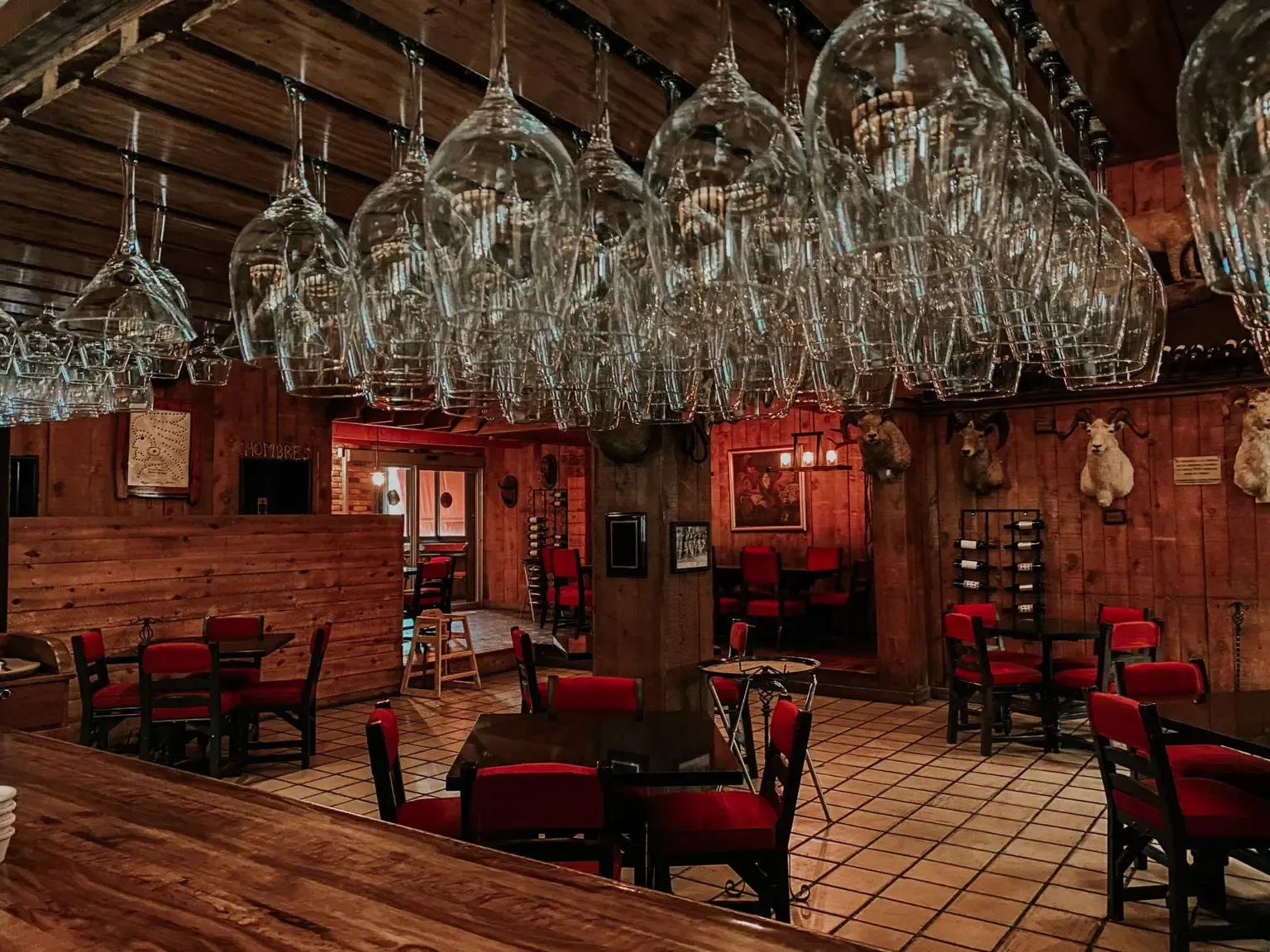 Copas de vino cuelgan sobre una barra rústica con sillas rojas y paneles de madera.