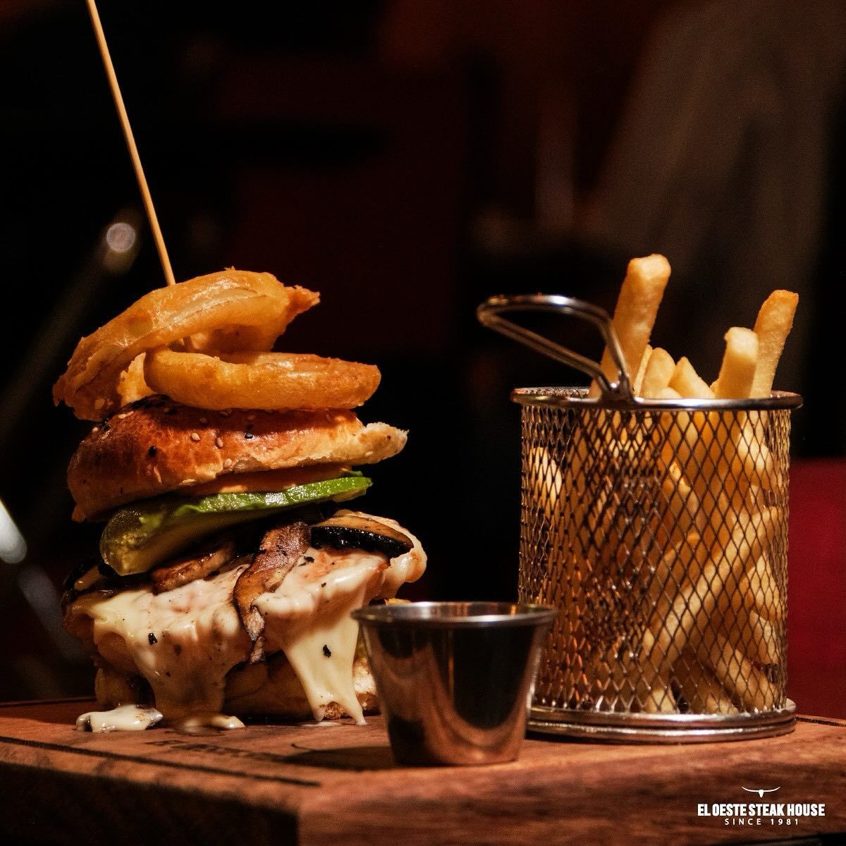 Hamburguesa con aros de cebolla y patatas fritas en una cesta metálica, servida en una tabla de madera.